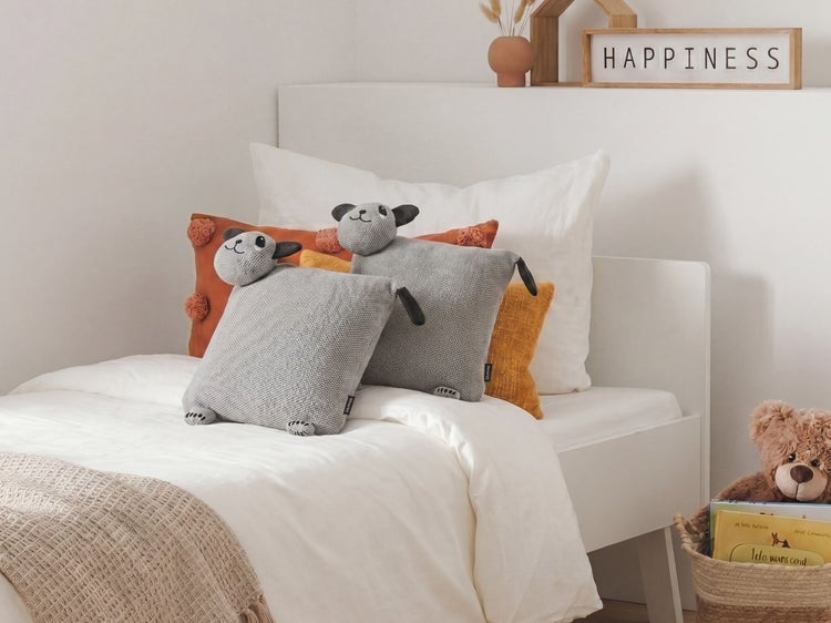 Helles Kinderzimmer mit weißem Bett, zwei grauen Strickkissen in Schaf-Form und verschiedenen Dekokissen.