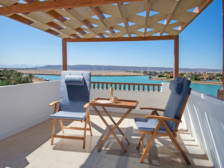 Zwei hölzerne Gartenstühle mit blauen Polstern und ein Klapptisch auf einer sonnigen Terrasse unter einer Pergola mit Blick aufs Wasser.