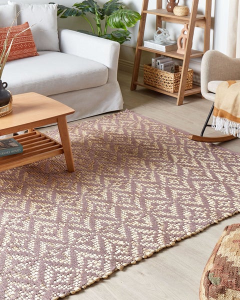 Gewebter Teppich mit lila und beigem geometrischem Muster in einem hellen Wohnzimmer mit hellem Sofa und hölzernem Couchtisch.