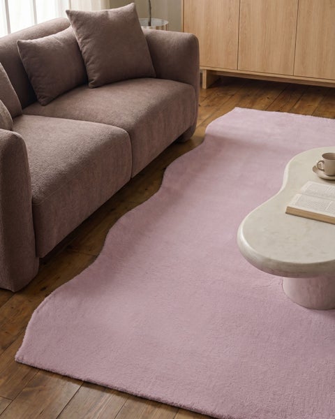 Flauschiger, unregelmäßig geformter Teppich in Rosa in einem gemütlich eingerichteten Wohnzimmer mit braunem Sofa und Couchtisch aus Stein.