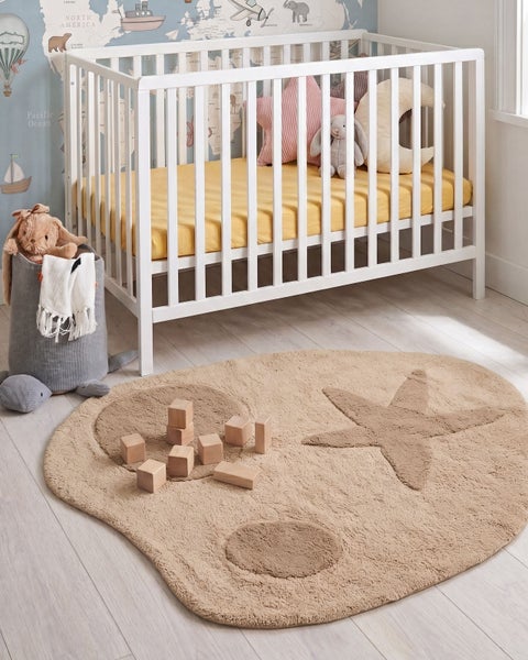Ein Kinderzimmer mit einem weissen Gitterbett, einem runden Spielteppich mit Sternmotiv und Holzbausteinen auf einem hellen Holzboden.