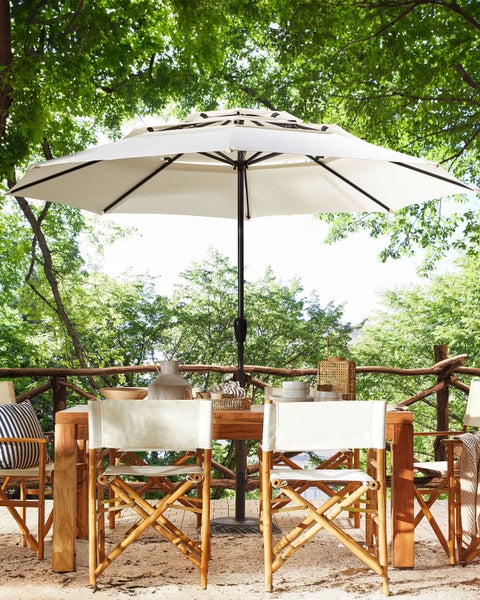 Großer weißer Marktschirm über einem hölzernen Esstisch mit Regiestühlen auf einer hellen Holzterrasse im Garten unter Bäumen.