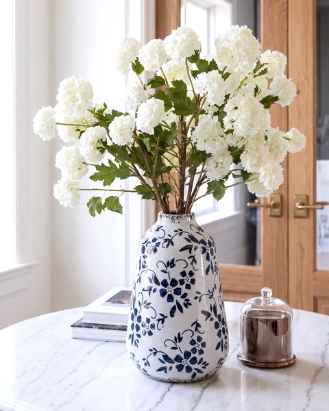 Dekorative Keramikvase mit Blumendekor und Strauß aus künstlichen Hortensien auf einem runden Marmortisch mit Kerze und Büchern als Wohnaccessoire