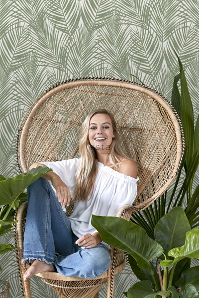 Frau sitzt in einem Rattansessel vor einer Wand mit Palmenblattmuster
