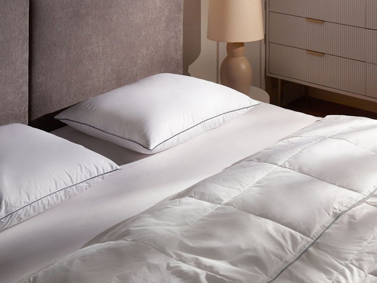 Helles Schlafzimmer mit weißer Bettwäsche, zwei Kopfkissen und einer gesteppten Bettdecke auf einem Bett mit grauem Kopfteil.