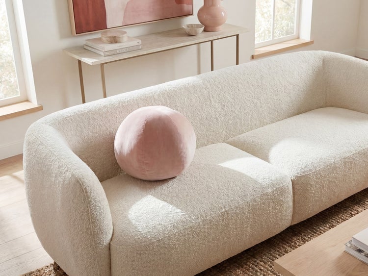 Texturiertes Bouclé-Sofa in einem hellen Wohnzimmer mit einem runden Samtkissen. Moderner Einrichtungsstil mit Konsole und Fenstern.