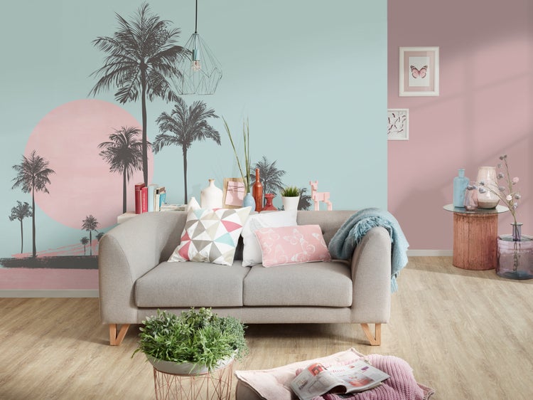 Modernes Wohnzimmer mit einem Zweisitzer-Sofa, Dekokissen, einer großen Fototapete mit Palmen und Sonnenuntergang sowie Holzboden.