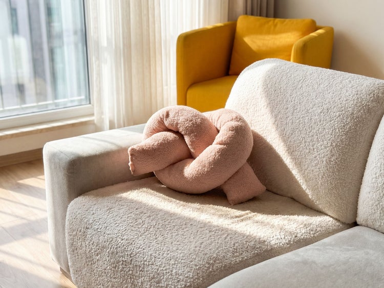 Knotenkissen aus Teddystoff auf einem Sofa in einem hellen Wohnzimmer mit Sessel und Vorhängen.