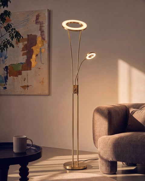 Moderne Stehleuchte aus Metall mit zwei ringfoermigen LED Leuchtkoepfen fuer Deckenfluter und Leselicht neben einem Sofa in einem Wohnraum.