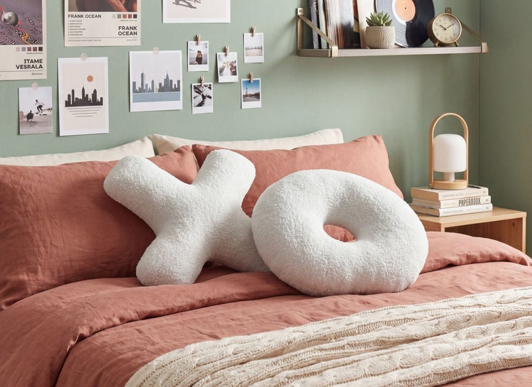 Helles, organisiertes Schlafzimmer mit zwei texturierten Plüschkissen in X- und O-Form auf einem Bett mit Leinenbettwäsche und Wanddekorationen.