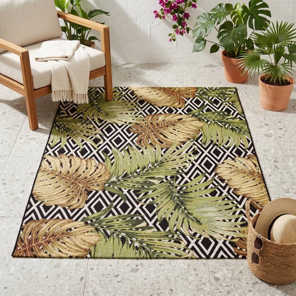 Outdoor-Teppich mit Monstera-Blattmuster und geometrischem Hintergrund auf einer hellen Terrasse mit Holzstuhl und Pflanzen.