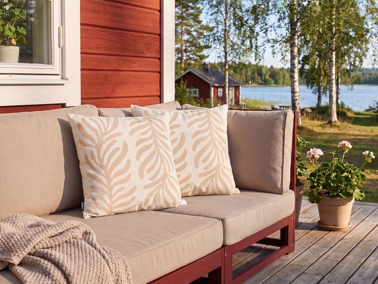 Gartensofa mit Kissen mit Blattmuster auf einer Holzterrasse vor einem Gartenhaus mit Blick auf einen See.