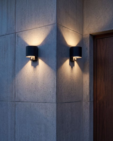 Zwei moderne Außenwandleuchten mit Bewegungsmelder an einer Betonfassade erzeugen Lichtkegel nach oben und unten.