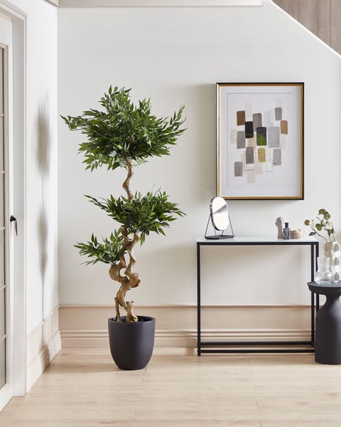 Dekorativer künstlicher Baum in einem schwarzen Topf neben einer Konsole mit Dekorationsgegenständen in einem hellen Raum.