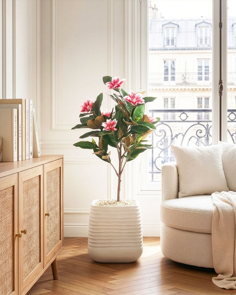 Künstliche Magnolie im gerippten Pflanzkübel in einem hellen, elegant eingerichteten Wohnzimmer mit Sessel und Holzkommode.