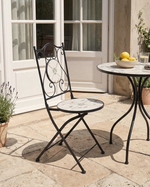 Klappbarer Mosaikstuhl und runder Mosaiktisch aus Metall auf einer Steinterrasse im mediterranen Stil vor einer hellen Flügeltür.