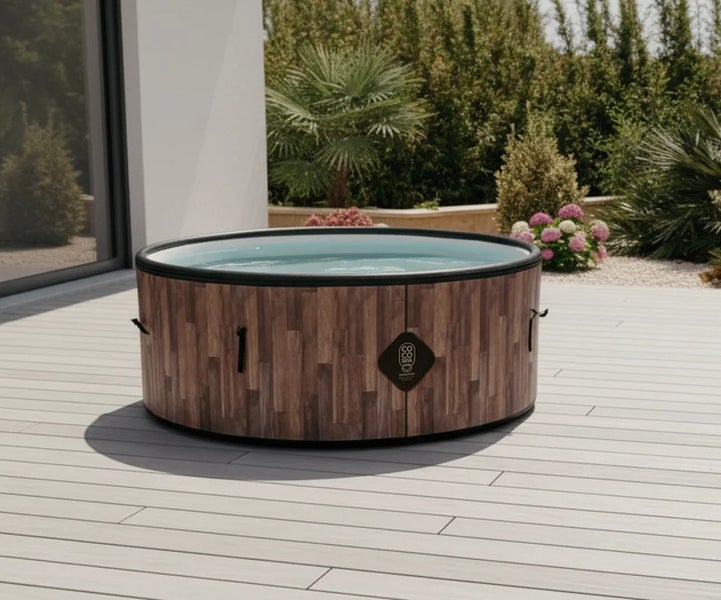 Runder Whirlpool mit vertikaler Holzoptik und COCO SPA Logo auf einer hellen Holzterrasse vor einer modernen Hauswand und grünem Garten.