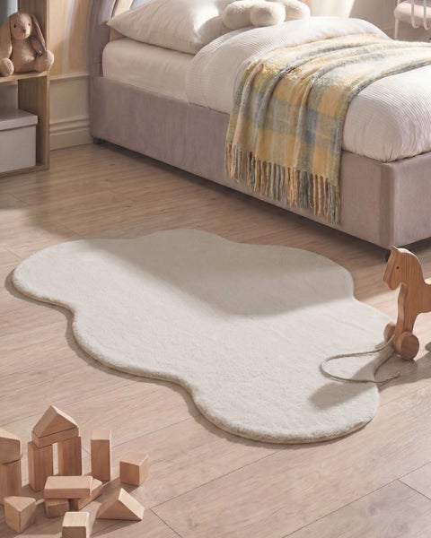 Weißer, wolkenförmiger Teppich in Felloptik auf einem Holzboden in einem hellen Kinderzimmer neben einem Bett und Holzspielzeug.