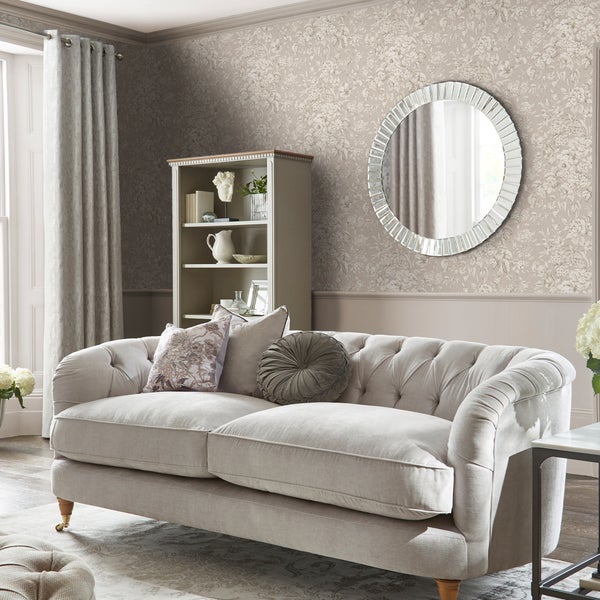 Ein elegantes Wohnzimmer mit einem beigen Chesterfield Sofa, einer geblümten Tapete, einem runden Wandspiegel und einem offenen Regal.