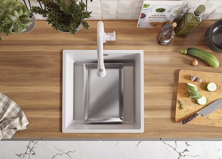 Spüle mit Abtropfsieb, Armatur, Schneidebrett mit Gemüse und Kochbuch auf einer Küchentheke.