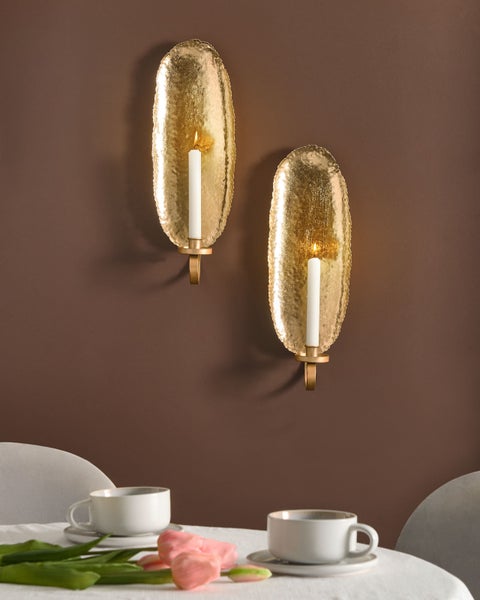 Zwei goldfarbene Wandkerzenhalter aus gehämmertem Metall mit weißen Kerzen an einer Wand über einem Tisch mit Kaffeetassen und Blumen.