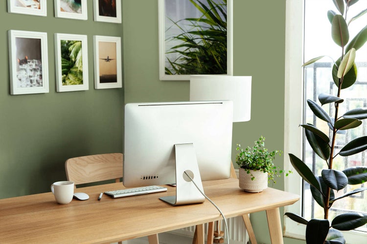 Modernes Homeoffice mit hellem Holztisch, Computer und Wandfarbe in Salbeigrün. Heller Arbeitsplatz mit Pflanzen und gerahmten Wandbildern.