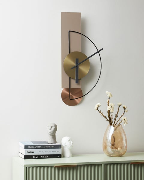 Moderne Wanduhr mit geometrischen Elementen aus Metall über einer Kommode mit Büchern, Kopf-Skulpturen und einer Vase mit Trockenblumen.