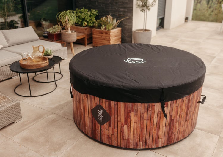 Runder Whirlpool in Holzoptik mit Abdeckung auf einer hellen Terrasse mit Loungemöbeln, Beistelltischen und Pflanzen in Holzkübeln.