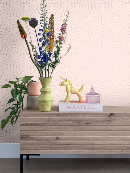 Dekoratives Stillleben mit Blumen, Pflanzen und Dekorationselementen auf einer Holzkommode vor einer gemusterten Tapete.