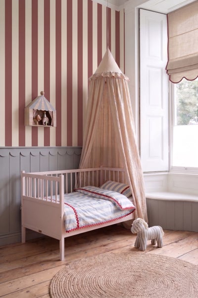 Kinderzimmer mit gestreifter Tapete, grauer Wandverkleidung, rosa Kinderbett mit Baldachin und rundem Juteteppich.