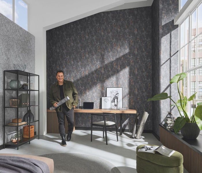 Ein modern eingerichtetes Zimmer mit einem Mann, der eine Rolle Tapete hält, einem Schreibtisch, einem Regal und einer Zimmerpflanze.