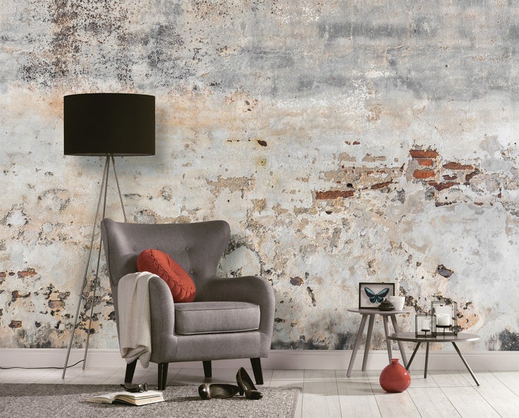 Vliestapete im Industrie-Design mit Beton- und Mauerwerk-Optik in einem Wohnzimmer mit grauem Sessel, schwarzer Stehlampe und Dekoration.
