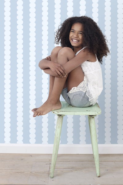 Ein Mädchen mit lockigem Haar sitzt lächelnd auf einem Hocker vor einer gemusterten Wand.