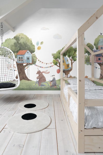 Kinderzimmer mit Holzbett, Hängesessel und runden Teppichen mit Punktmuster vor einer Wandtapete mit Waldmotiv.