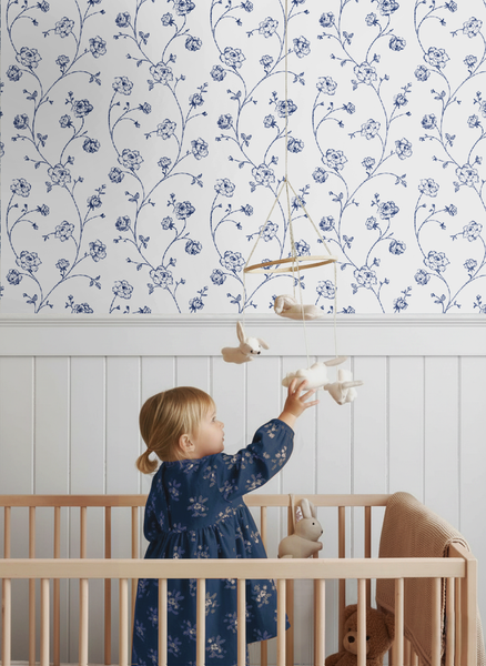 Kinderzimmer mit blumiger Tapete in Blau und Weiß sowie weißer Wandverkleidung aus Holz. Ein Kind im Gitterbett greift nach einem Mobile.