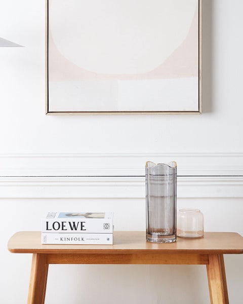 Dekorative Szene mit Bücherstapel, grauer Vase und kleiner Glasdose auf einem Holztisch unter einem gerahmten Bild.