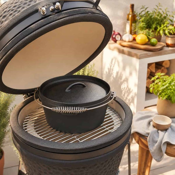 Gusseisen Dutch Oven in einem offenen Keramikgrill auf einer Terrasse mit Kräutern und Küchenutensilien im Hintergrund