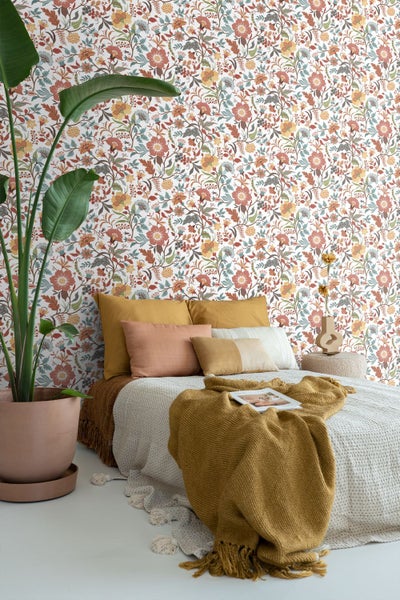 Gemütliches Schlafzimmer mit detailreicher Blumentapete in warmen Tönen an der Wand, einem Bett mit Kissen und einer großen Zimmerpflanze.