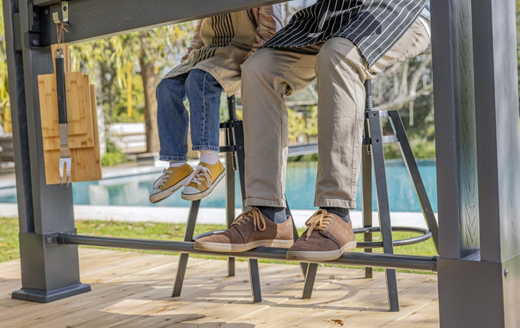 Zwei Personen sitzen auf Barhockern an einem Bartisch im Freien. An dem Bartisch hängen Schneidebretter und eine Grillgabel.