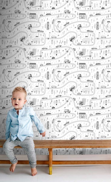 Kindertapete mit Muster von Autos, Straßen und Häusern in einem Kinderzimmer, ein kleiner Junge sitzt auf einer Holzbank davor.