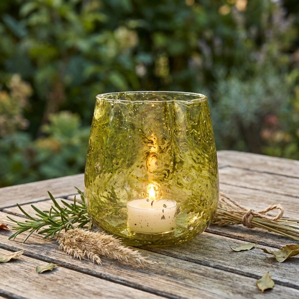 Ein bauchiges Windlicht aus strukturiertem Glas auf einem Holztisch mit einem brennenden Teelicht im Freien.
