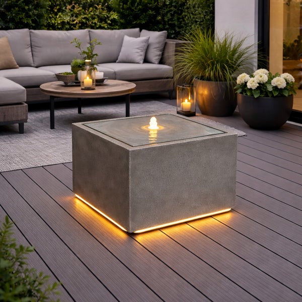 Moderne Terrasse mit quadratischem Gartenbrunnen in Beton-Optik, Wasserspiel und LED-Beleuchtung, umgeben von Lounge-Möbeln und Pflanzen.