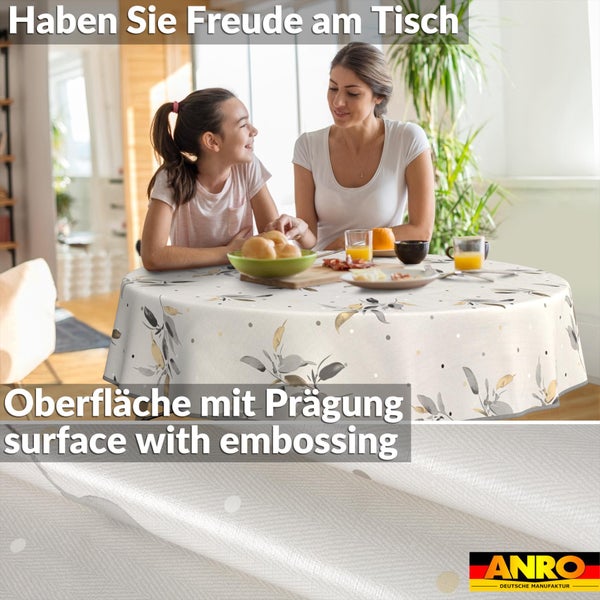 Runde Tischdecke mit geprägter Oberfläche und Blattmuster in einer hellen Esszimmerszene mit Frau und Kind. ANRO DEUTSCHE MANUFAKTUR Logo.