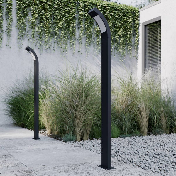 Moderne Außenwegeleuchten in einem hellen Garten mit Ziergräsern, Kieselsteinen und einem Betonweg vor einer mit Efeu bewachsenen Wand.