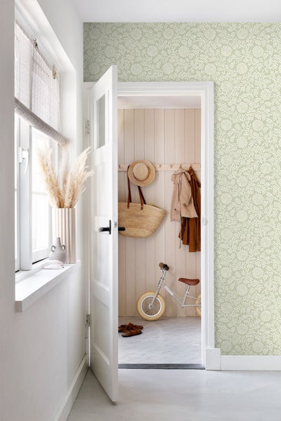 Hell gestalteter Flur mit floral gemusterter Tapete, weißer Tür und Blick in einen Raum mit Holzpaneelen und Kinderlaufrad.