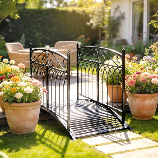Kleine dekorative Gartenbrücke aus Metall mit bogenförmigem Geländer und Ornamenten in einem hellen Garten.