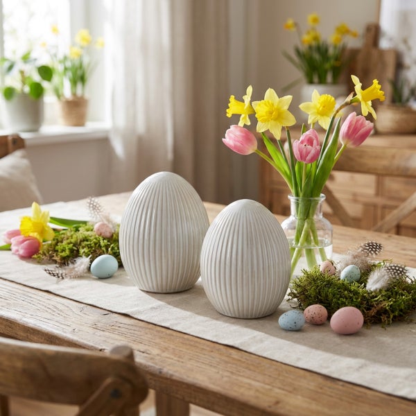 Osterdekoration auf einem Holztisch mit zwei gerillten Eiern aus Keramik, einer Glasvase mit Frühlingsblumen, Moos und kleinen Ostereiern.