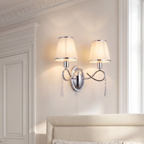 Zweiflammige Wandleuchte aus Chrom mit plissierten Stoffschirmen und Kristallanhängern über einem Bettkopfteil in einem hellen Zimmer.