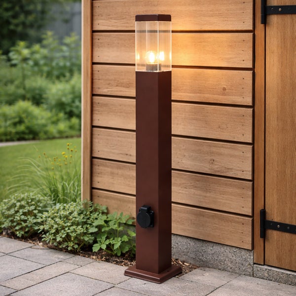 Moderne Außenstandleuchte in Braun mit integrierter Steckdose und Glasaufsatz mit brennendem Leuchtmittel vor einer Holzwand im Garten.