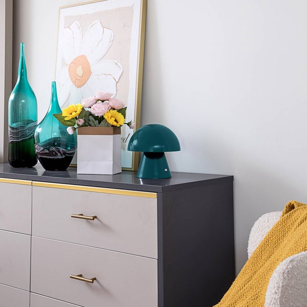 Moderne graue Kommode mit Goldgriffen. Auf der Kommode stehen dekorative Glasvasen, ein Blumenstrauß und eine pilzförmige Tischleuchte.